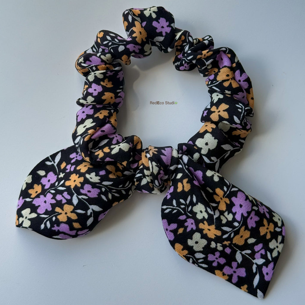 Angled view of the Violet Meadow mini pure silk scrunchie, showing its soft gathers and bow tails in a black floral print